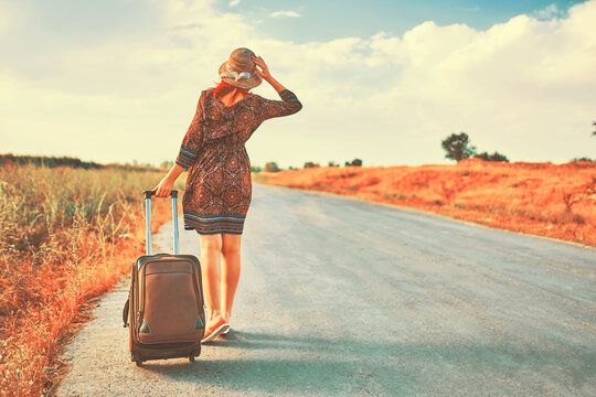 Very awesome female traveler in summer dress and hat hitchhiking with big bag at fall. Cute woman walking on the road with baggage. Tourism concept