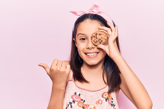 Beautiful child girl holding cookie pointing thumb up to the side smiling happy with open mouth