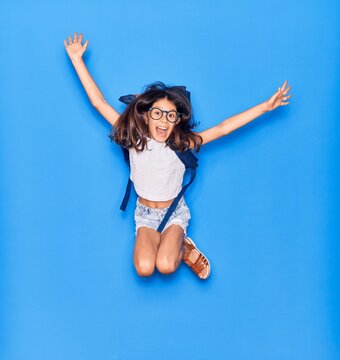Adorable hispanic student child girl wearing glasses and backpack with open mouth. Jumping excited with arms opened over isolated blue background