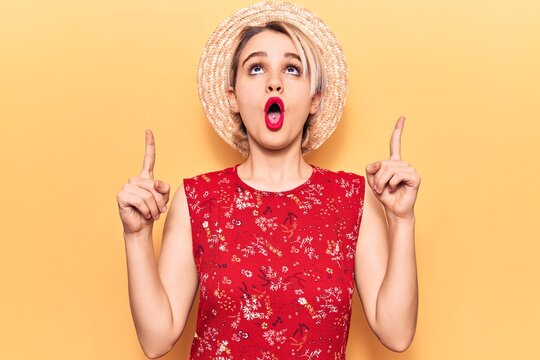 Young beautiful blonde woman wearing summer hat amazed and surprised looking up and pointing with fingers and raised arms.