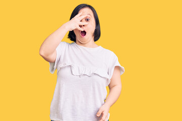 Brunette woman with down syndrome wearing casual white tshirt peeking in shock covering face and eyes with hand, looking through fingers with embarrassed expression.