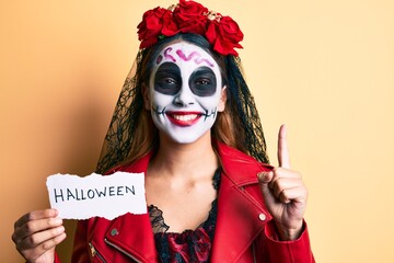 Woman wearing day of the dead costume holding halloween paper smiling with an idea or question pointing finger with happy face, number one