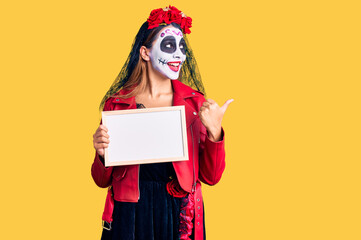 Woman wearing day of the dead costume holding empty white chalkboard pointing thumb up to the side smiling happy with open mouth