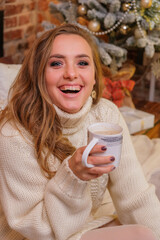 Portrait of young woman with cup of cocoa on bed at home. Minimalist christmas cozy decor on the background. Blonde, cute, woman in white sweater and warm scandinavian socks. New Year’s Eve mood.