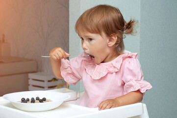 Little kid girl eating her morning porridge oatmeal with blueberries herself, sitting in baby chair in kitchen