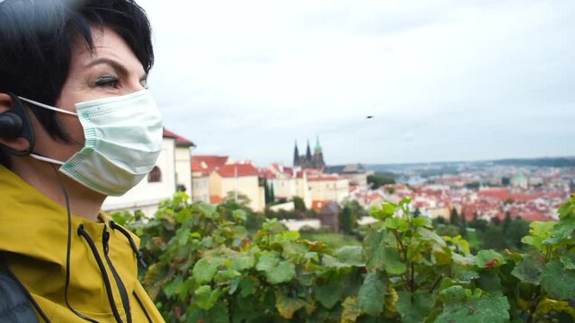 Audio guide tour of a European city during a next wave of viral pandemic. A woman in a protective mask listens to the sights with a earpiece, looks at the horizon. Autumn peak of the disease.