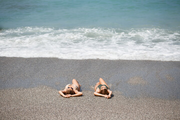 Fototapeta na wymiar Two girls girlfriend are sitting on the sea sandy shore and the waves soaked them in bathing suits on a sunny warm day