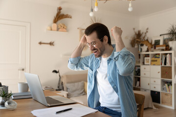 Overjoyed young man raising arms in air, making yes gesture celebrating online lottery win or personal professional success at home office, received email notification with good news on computer.