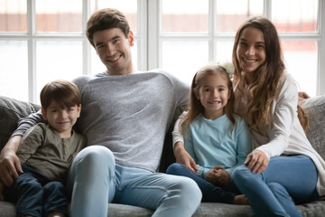 Portrait of smiling young Caucasian family with two small kids sit relax on sofa in living room together. Happy parents rest on couch on weekend at home with little children, show love and care.