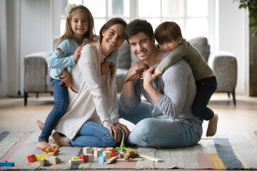 Portrait of happy young Caucasian family with small kids sit on floor in living room have fun on weekend together. Smiling parents with little children play at home. Tenancy, relocation concept.