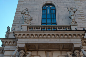 Ostrovsky Square 2a, Saint Petersburg, Russia, 11.10.2020. Ancient sculpture stands on balcony of facade in wall of gray building with masonry. Blue sky reflection in window