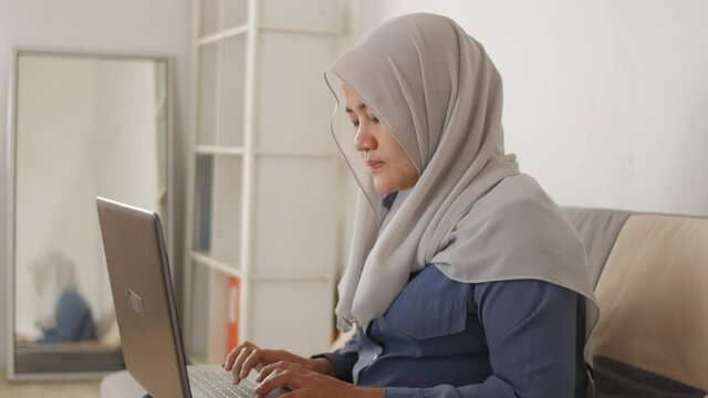 Asian muslim woman wearing hijab smiling while typing on her laptop, work from home concept. Young successful female businesswoman or entrepreneur working at home