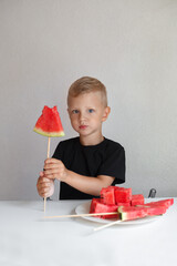 a boy in a black T-shirt holds a slice of ripe watermelon on a wooden stick and looks at the camera, next to there is a plate with square slices of watermelon. Image with selective focus