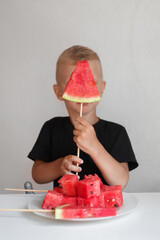 a boy in a black T-shirt holds a slice of ripe watermelon on a wooden stick, next to it is a plate with square slices of watermelon. Image with selective focus