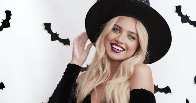 Halloween costume. Portrait of blonde woman looking at camera and smiling, touching her witch hat