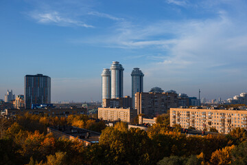 Fototapeta premium Urban landscape of new Moscow. Urban area with old and new housing during sunny day with foggy clouds and blue sky. In foreground are autumncolored treetops. Contrasty scene in architecture and light.