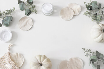Decorative cosmetics frame. Organic cotton reusable round pads for make up removal. Eucalyptus branches, pumpkins.and moisturizer on white table background. Zero waste concept. Flat lay, top view.