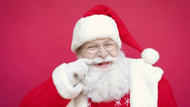 Close up portrait of funny happy old bearded Santa Claus face wearing hat, glasses, touching mustache, looking at camera, laughing, standing on Merry Christmas, Happy New Year red background.