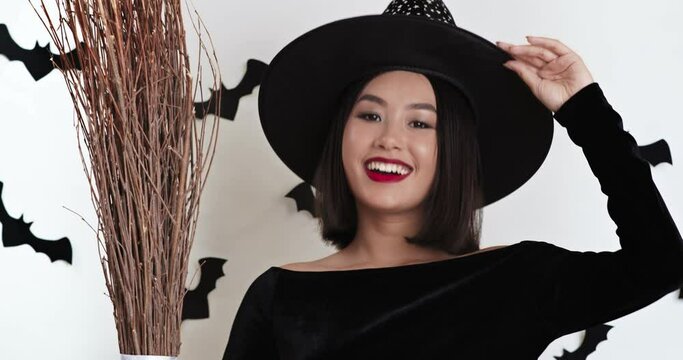 Cheerful asian woman wizard wearing black costume with broom, smiling at camera and waving hello, white background