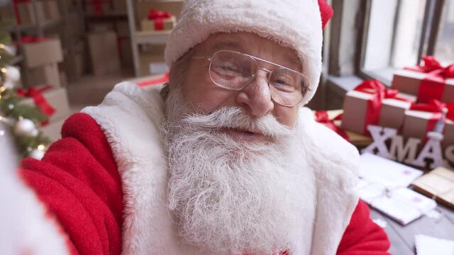 Happy old bearded Santa Claus wearing costume and hat holding phone waving hand video calling in zoom, recording video Merry Christmas greeting or shooting vlog standing in workshop, face camera view.