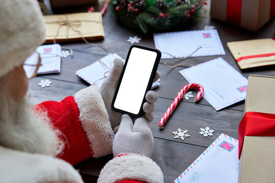 Over shoulder view of Santa Claus wearing costume holding smartphone with white blank display screen using mobile app sitting at workshop table on Merry Christmas eve. Ecommerce application ad closeup