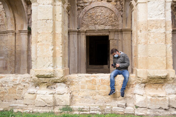 Fototapeta na wymiar Joven sentado en un muro de una iglesia en ruinas con mascarilla higiénica y mirando su cámara de fotos