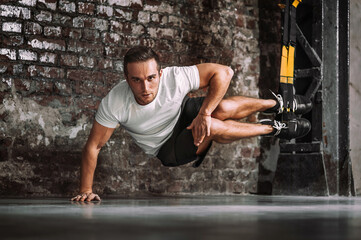 Serious sportsman with legs on TRX ropes looking at camera during intense suspension workout against rough brick wall