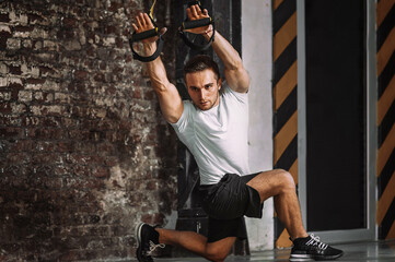 Fit young man in sportswear lunging and raising arms on suspension ropes during intense workout in grungy gym