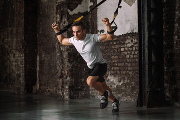 Fototapeta na wymiar Full body strong male athlete in sportswear clenching fists and sprinting forward while exercising with TRX ropes during intense training in grungy gym