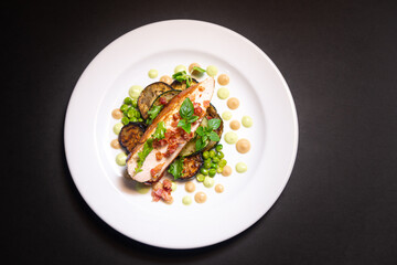 Turkey breast meat with fried pieces of bacon sits on baked eggplant  and green peas aside, young and small basil leaves and cauliflower puree and pea puree. On a white plate and a black background.
