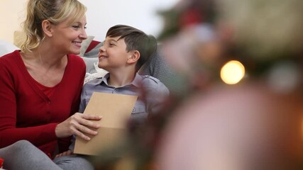 Merry christmas, happy family at home, son thanks for christmas gift giving a kiss to mom near the illuminanted and decorated tree, sitting in living room - Powered by Adobe