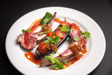 Lamb ribs with baked eggplant, cherry tomato wedges, caramelized onions, small basil leaves, micro greens, with red sauce on a white plate on a dark black background. Side view.