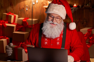 Happy old Santa Claus wearing hat holding gift box using laptop computer sitting at workshop home...