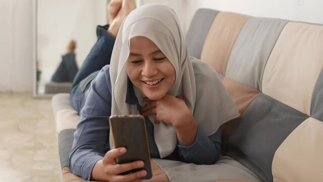 Beautiful Asian muslim lady doing selfie portrait on phone or doing video call while sitting on sofa, happy smiling cheerful expression
