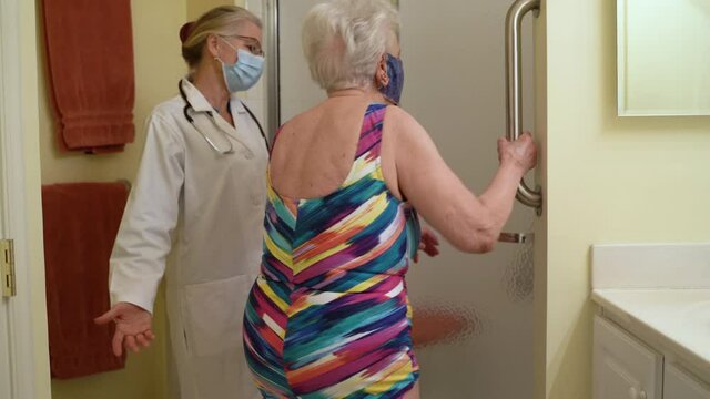 Wearing face masks, woman doctor caregiver helps elderly woman into shower with grab bar retirement home senior health assistance.
