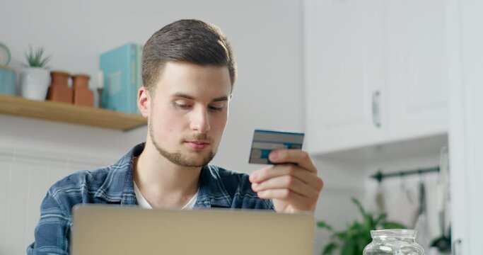 Happy Young Man is Paying Online by Credit Card, making Purchase at Home in Kitchen in Web Store using Laptop. E-banking and E-commerce app service Concept. Person Enjoys Shopping via Internet.