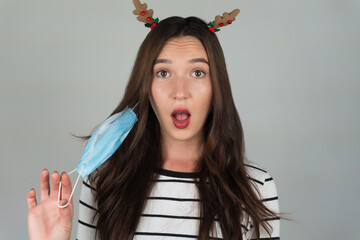 Beautiful young brunette with a medical protective mask on a gray background. A woman poses with a Christmas decoration in her hair. Celebrating the New Year in the midst of a pandemic.
