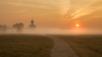 A silhouette of a Russian Orthodox church above a misty field backlit by the rising sun. A stone-cobbled road leads to the church. No people.