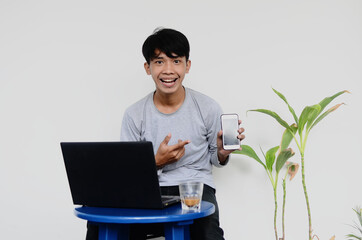 young Asian man who sits in front of the laptop while showing his cell phone