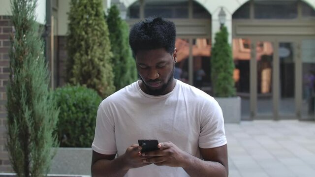 Portrait of young African American man is texting message and then talking on her smartphoneis. Stylish brunet in a white T shirt on the background of the street outdoors. Slow motion. Close up.