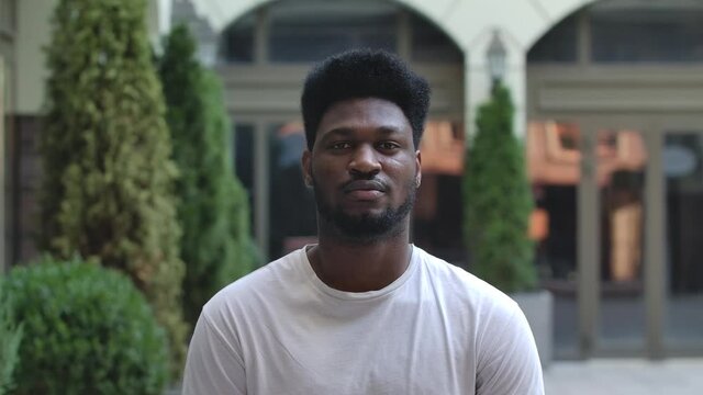 Portrait of young African American man looking at the camera. Stylish brunet in a white T shirt on the background of the street outdoors. Slow motion. Close up.