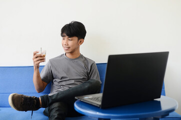 Asian youth sitting on the sofa in front of the laptop while drinking coffee