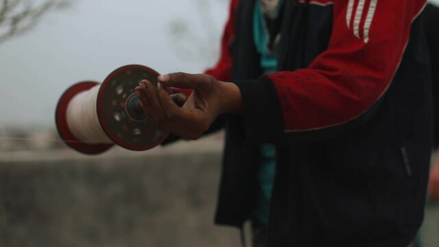 close up footage of male hands holding and speed rolling thread from a spool while controlling a kite flying high in sky. day time outdoor rooftop footage from rural delhi, india.