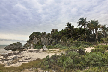 Fototapeta na wymiar Beach on the Caribbean coast. A small sandy area surrounded by rocks and palm trees. At the top are the ruins of the ancient Mayan city of Tulum. Cloudy sky. Mexico.
