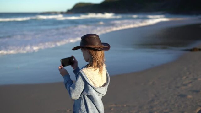 A boy with a hat makes photoes of the ocean waves on a sandy beach.