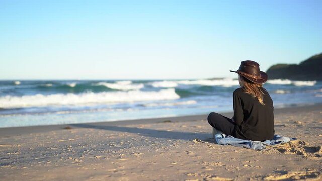 A boy in dark clothes sits on a sand beach looking at ocean waves.