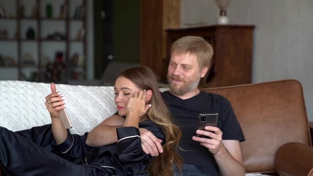A man and a woman in love are hugging and watching a funny video or picture on the smartphone display at home while lying on the sofa in an embrace.