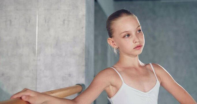Pretty girl ballerina with bun in white costume leans on wooden barre posing at large mirror in dance studio closeup slow motion