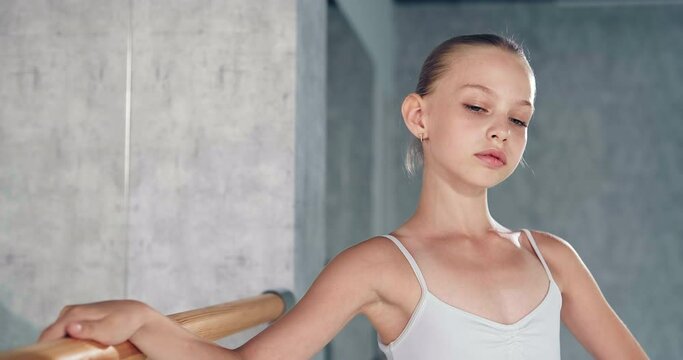 Graceful girl ballerina with bun in white costume stands near wooden barre posing as actress in dance studio closeup slow motion
