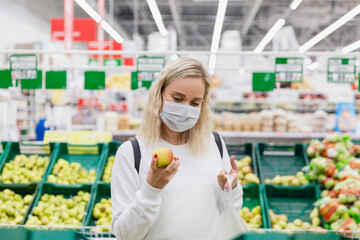 Young woman in a medical mask chooses apples in a supermarket. Healthy eating. Coronavirus pandemic.